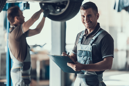 Mechanics Checking Wheel Bearings in Car Workshop. Two Caucasian Adult Technician Master in Uniform Repairing Car. Repairman Looking at Camera Filling Clipboard of Service Order. Auto Service Conceptの写真素材