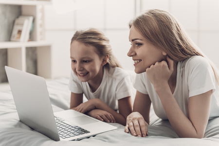 Young Mother and Little Doughter using Laptop. Beautiful Smiling Woman spending time with Cute Adorable Daughter. Mom and Child lying on Bed and using Laptop at Home. Family and Motherhood Conceptの写真素材