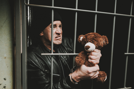 A Man In Handcuffs Is Behind Bars In A Police Station. Jailed Suspect. Bandit In Leather Jacket. Successful Arrest. Commit A Crime. Guilt Negation. Close-up Criminal. Plush Toy Holding.の写真素材