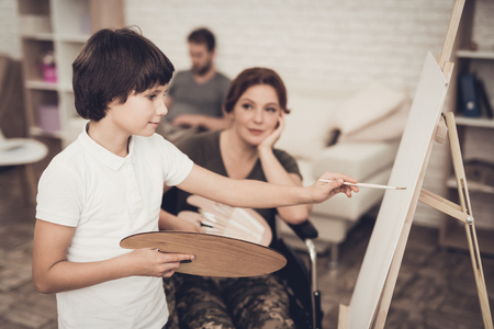 Disabled Soldier's Son. Picture Drawing Concept. After War. Camouflage Uniform. Paralyzed Woman. Child With Tassel. Home Leisure. Return From Army. Family Background. Young Artist.の写真素材