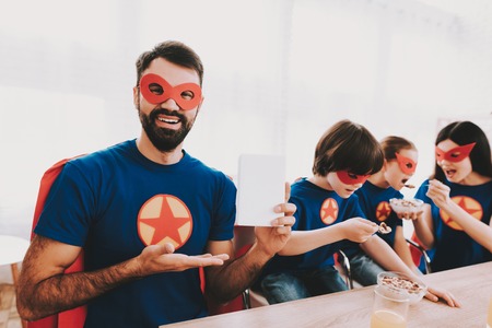Young Family In Superhero Suits. Dinner Concept. Masks And Raincoats. Bright Room. Resting Together....