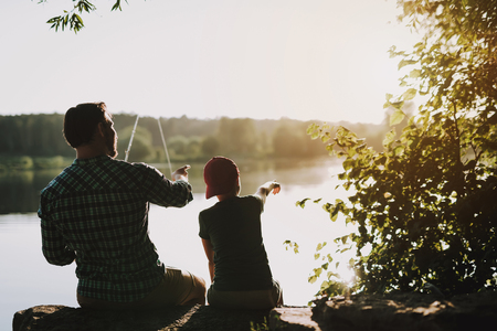 Young Father with Son in Cap Fishing on River in Summer. Relaxing Outdoor. Bearded Young Man. Men near Lake. Fishing Rod in Hands. Sports in Summer. Weekend on River. Young Fisherman.の写真素材