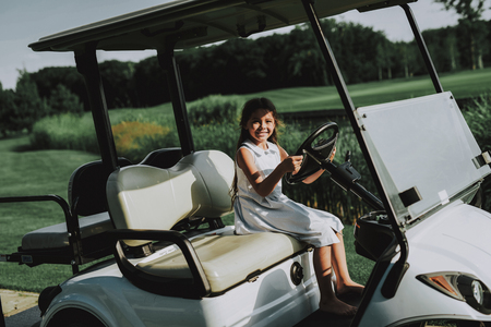 Little Girl in White Dress in Cart on Golf Field. Smiling Girl. White Dress. Healthy Lifestyle Concept. Golf Club. Sports in Summer. Vehicle on Field. Family Holiday. Outdoor Fun in Summer.の写真素材
