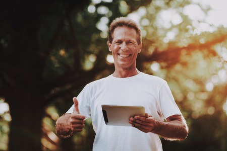 Man Standing in Summer Park and Using Tablet. Sport and Healthcare Concept. Young Man with Gadget. Summer Park. Fun Outdoor. Using Digital Device. Trainer in Summer Park. Smiling Young Coach.の写真素材