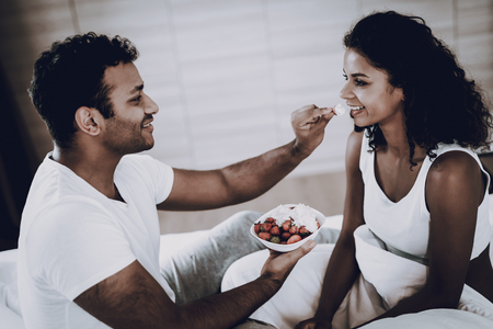 Man Is Feeding His Girlfriend With A Strawberry. Weekend Concept. Chilling On Sofa. Holiday Resting. Smiling Together. Having Fun. Romantic Date. Afro American Couple In The Bedroom.の写真素材