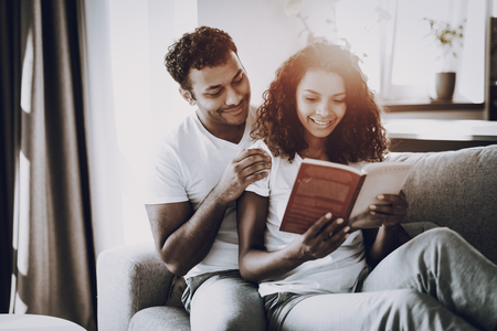 Man Is Hanging Book Reading Girlfriend On Couch. Morning Leisure. Have A Break. Home Clothes. Holiday Resting. Smiling Together. Happy Young Afro American Couple. Sunny Day. Love Each Other.の写真素材