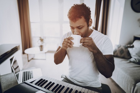 Young Man Drinks Coffee Near Synthesizer At Home. Happy Songwriter. Morning Leisure. Digital Format...