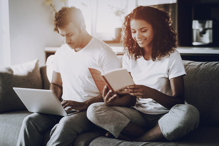 Couple With Laptop And Book Sitting On The Couch. Morning Leisure. Have A Break. Look On The Screen. Chilling On Sofa. Home Clothes. Holiday Resting. Smiling Together. Hands On Keyboard.の写真素材