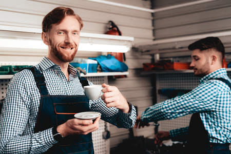 Two Young Cheerful Mechanics Are Drinking Coffee On A Service Station. Relaxing Concept. Having A Break. Cup And Saucer. Smiling Colleagues. Professional Uniform. Working In A Garage.の写真素材