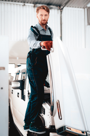 Red-haired Mechanic Stands On Wheel. Open Hood.Professional Uniform. Confident Engineering Specialist Stare. Protective Gloves. Working In The Garage. Young Repairman Posing Concept. Big SUV.の写真素材