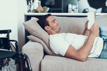 Man Lying on Couch with Tablet near Wheelchair. Cheerful Handsome Young Smiling Disabled Person...