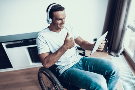 Disabled Man in Headphones Communicate Via Tablet. Handsome Smiling Person Wears T-Shirt and Blue Jeans Sits in Wheelchair Browsing Internet and Talking Via Video and Showing Thumbs up.の写真素材
