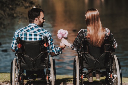 Couple of Disabled People on Wheelchairs on Date in Park. Young Man with Flowers. Woman on Wheelchair. Date in Summer Park. Romantic Relationship. Recovery and Healthcare Concepts. Happy Couple.の写真素材