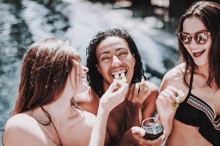 Young Smiling Women Drinking Wine at Poolside. Group of Young Happy People at Poolside holding Glasses of Wine and Eating Grapes. Happy Friends Enoying Pool Party. Summer Vacation Conceptの写真素材