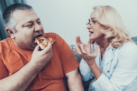 Upset Doctor in Coat near Fat Man with Pizza. Man with Bulimia. Unhealthy Lifestyle Concept. Sitting Man. Eating Pizza. Woman in White Coat. Patient with Stomach. Man with Overweight.の写真素材
