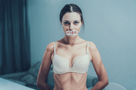 Anorexic Woman in White Bra with Tape on Mouth. Girl with Anorexia. Weigth Problem Concept. Woman in Bra. Girl on Diet. Sad Girl. Starvation Concept. Woman in Gray Room. Depressed Girl.の写真素材