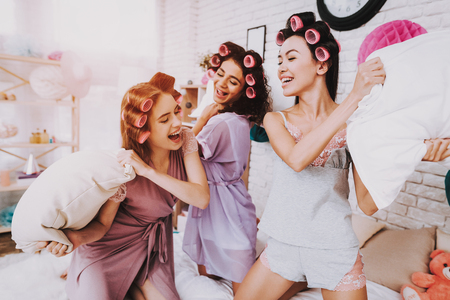 Fight Pillows. Home Party. Women in Bathrobes. Pillows on White Bed. Bright Room. White Interior. Beautiful International Woman. Smiling Girls. Pillow in Hand. Funny People. Women with Pink Curlers.の写真素材