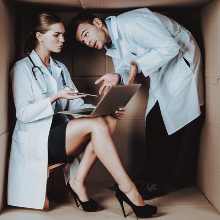 Two Young Doctors in White Coats in Cardboard Box. Young Woman in White Coat. Life in Little Cardboard Box. Uncomfortable Life. Personal Spase Concepts. Medical Equipment. Doctor witn Laptop.の写真素材