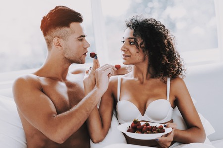 Happy Romantic Couple Eating Strawberrys in Bed. Young Beautiful Couple in Underwear Sitting on Bed with Plate of Fresh Berries. Handsome Man and Attractive Woman in Love. Relationship and Loveの写真素材