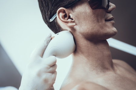 Young Man in Spa Salon for Laser Hair Removal. Guy in Beauty Salon. Modern Cosmetology. Proffesional Cosmetologist. Doctor with Laser Epilator. Men's Beauty Concept. Man in Glasses.の写真素材