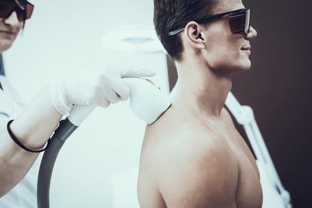 Young Man in Spa Salon for Laser Hair Removal. Guy in Beauty Salon. Modern Cosmetology. Proffesional Cosmetologist. Doctor with Laser Epilator. Men's Beauty Concept. Man in Glasses.の写真素材