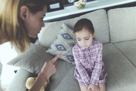 Mother Scolding Upset Pretty Child Indoors. Caucasian Girl Sitting on Comfortable Couch in Modern Living Room Feels Guilty. Swearing Mother Point with Finger at Daughter. Family Relationship Conceptの写真素材