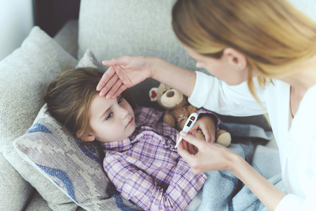 Careful Mother Checking Sick Child Temperature. Attentive Woman Sits Near Ill Little Daughter. Pretty Girl has Cold and Fever Lying on Sofa under Blanket or Plaid. Careful Mom Holding Thermometerの写真素材