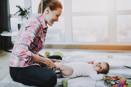 Happy Mother Plays with Cute Baby Lying on Carpet. Smiling Mom Wears Casual Clothes Resting with Sweet Beautiful Caucasian Toddler near Large Panoramic Window in Living Room. Childhood Conceptの写真素材