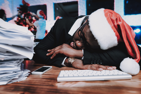 Tired Man in Cap Working in Office on New Year Eve. Christmas Tree in Office. Working Businessman.の写真素材