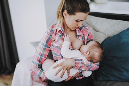 Young Mother Breast Feeding Little Baby Indoors. Portrait of Happy ?aucasian mother Sitting on Couch in Living Room and Holding Cute Adorable Child on Knees. Parenthood and Childhood Conceptの写真素材