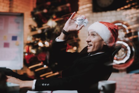 Businessman in Red Cap in Office on New Year Eve. Christmas Tree in Office. Business Concept. Man in Black Suit. Celebrating of New Year. Santa Claus. Happy Man. Smiling Businessman.の写真素材