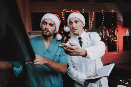 Two Doctors in Red Caps with X-ray Scan in Office. New Year Eve. Christmas Tree in Office. Computer on Desk. Man in Uniform. Celebrating of New Year. Santa Claus Cap. Medical Dispute.の写真素材