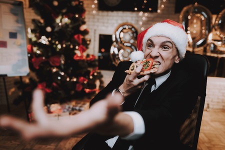 Man Watching Movie on Computer and Eating Burger. Christmas Tree in Office. Laptop on Desk. Business Concept. Man in Black Suit. Celebrating of New Year. Using Digital Device. Red Cap.の写真素材