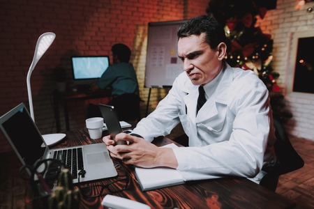 Tired Doctor with Tablet in Office on New Year Eve. Christmas Tree in Office. Computer on Desk. Healthcare Concept. Man in White Coat. Celebrating of New Year. Using Digital Device.の写真素材