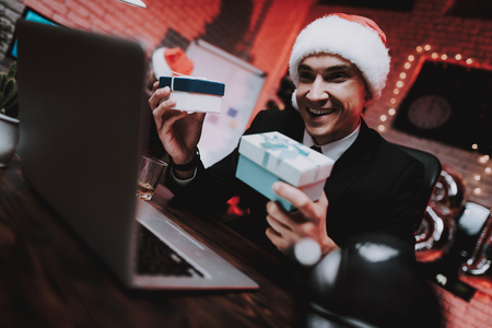 Businessman in Red Cap in Office on New Year Eve. Christmas Tree in Office. Laptop on Desk. Business Concept. Man in Black Suit. Celebrating of New Year. Using Digital Device. Gift Box.の写真素材