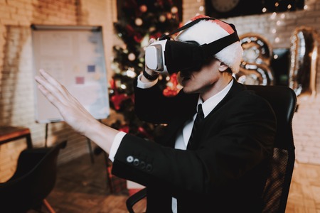 Man using VR-glasses in Office on New Year Eve. Christmas Tree in Office. Laptop on Desk. Man in Black Suit. Celebrating of New Year. Using Digital Devices. Red Cap. Man in Office.の写真素材