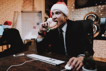 Businessman Playing on Computer and Eating Burger. Christmas Tree in Office. Laptop on Desk. Business Concept. Man in Black Suit. Celebrating of New Year. Using Digital Device. Red Cap.の写真素材
