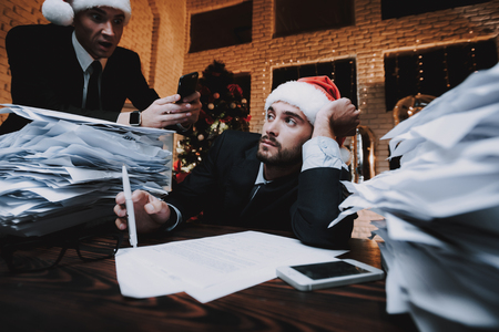 Tired Man Working with Documents on New Year Eve. Christmas Tree in Office. Business Concept. Man in Black Suit. Celebrating of New Year. Using Digital Device. Young Man. Tired Businessman.の写真素材