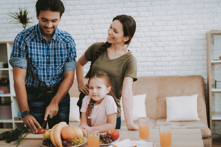Father Prepares Breakfast. Family Breakfast. Family at Table. Fruit on Plate. Sandwich on Plate. White Plate. Juice in Glass. Happy family. Smiling People. Vegetables on Board. White Interior.の写真素材