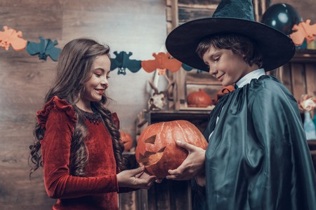 Adorable Little Children in Halloween Costumes. Cute Smiling Kids wearing Scary Halloween Costumes Standing holding Orange Carved Pumpkin. Cute Girl and Boy Celebrating Halloweenの写真素材
