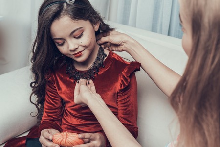 Beautiful Woman putting Black Necklace on Little Girl. Beautiful Mother helping Cute Smiling Daughter with Costume for Halloween Party. Celebration of Halloween. Family and Childhood Conceptの写真素材