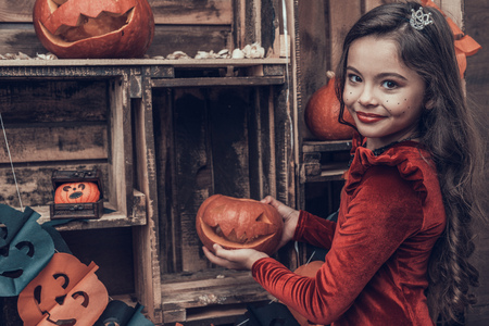 Cute Girl in Halloween Costume with Carved Pumpkin. Portrait of Cute Smiling Girl wearing Scary Halloween Costume holding Orange Carved Pumpkin. Celebration of Halloween. Traditional Seasonal Holidaysの写真素材