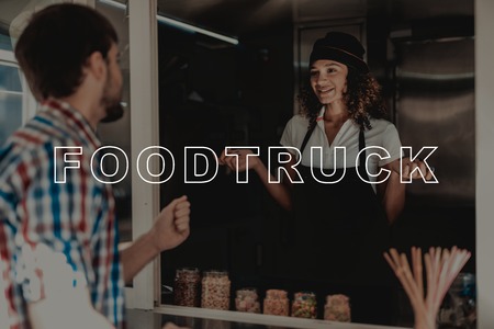 Man In A Plaid Shirt Buys Coffee In A Food Truck. Black Beard. Ready To Work. Fast Eating Place. Service Personal. Opened Window. Professional Apron Uniform. Sunny Day. Girl Seller With Curly Hair.の写真素材