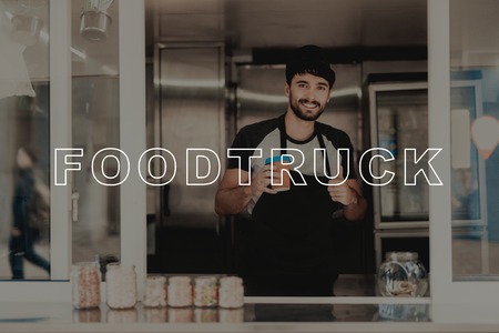 The Seller With Coffee Stands In The Food Truck. Black Beard. Ready To Work. Waiting For Clients. Fast Eating Place. Service Personal. Opened Window. Apron Work Uniform. Sunny Day.の写真素材