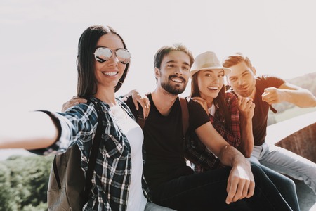 Young Smiling People Sitting in Park taking Selfie. Group of Young Friends with Backpacks Sitting Together taking Selfie by Using Smartphone. Travelers Enjoying Summer. People and Friendship Conceptの写真素材