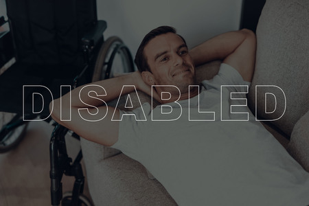 Disabled Man. Man Lying on Sofa. Wheelchair on Background of Man. Man is Relaxed. Adult Brunette Man. Man Happy and Smiling. Man Located in the Living Room. Daytime. Home Interior.の写真素材
