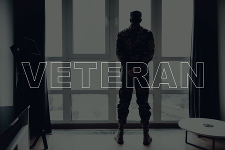 Military Veteran. Man Standing in Front of Window. Large Panoramic Window. Man is Soldier. Soldier in Military Uniform. Man Looking into Window. Man Located in the Living Room. Back View.の写真素材