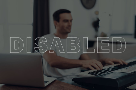 Disabled Man in a Wheelchair. Man Sitting at Table. Man Playing a Music on Synthesizer. Open Laptop on Wooden Table. Adult Brunette Man. Man is Located in the Living Room. Blur View.の写真素材