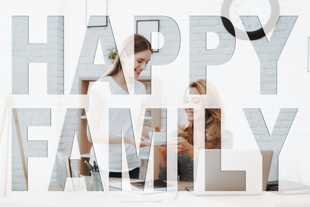 Happy Family. Mother Sitting at Table. Daughter Standing near Mother. Daughter Giving Tea Cup to Mother. Mother Taking Tea Cup. People Happy and Smiling. People Located in Home Apartments.の写真素材