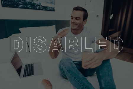 Disabled Man. Man is Sitting on Bed. Laptop Computer Lying on Bed. Smartphone and Notebook Lying on Bed. Man is Happy and Smiling. Man Eating a Cake. Man Drinking Juice in Cup. Man Located in Bedroom.の写真素材